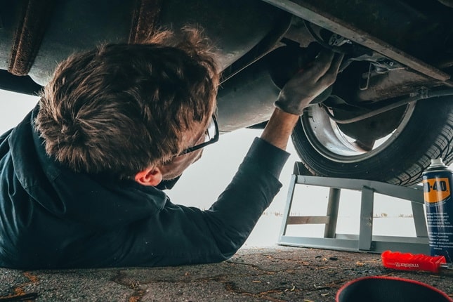 Man lying under a car performing maintenance work with tools and WD-40 can nearby.